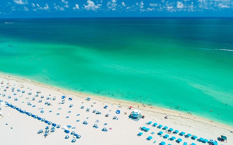 Aerial view of a resort's private clear water beach lined with lounge chairs and umbrellas in Florida
