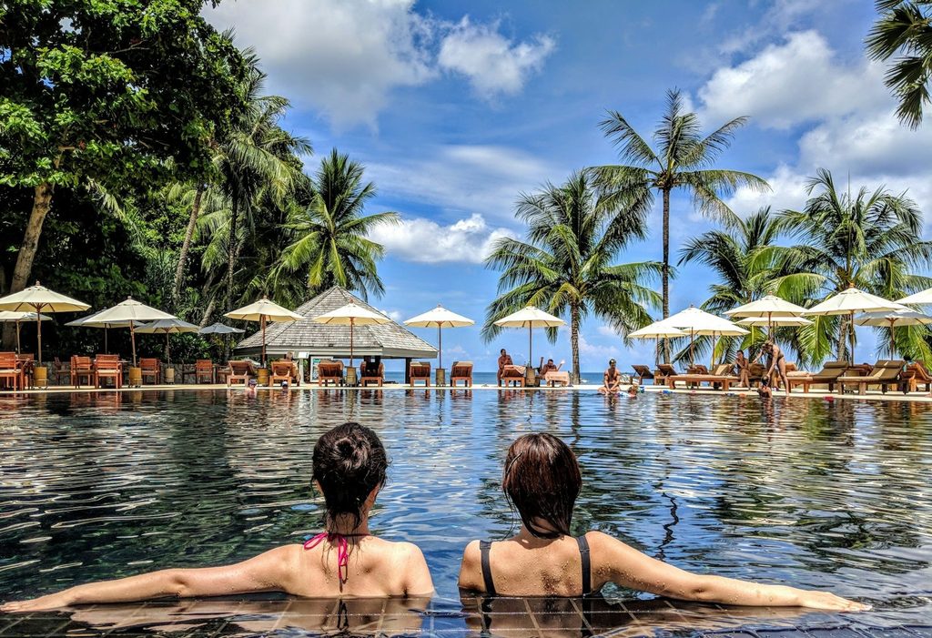 Two women relaxing at the edge of a resort infinity pool lined with palm trees
